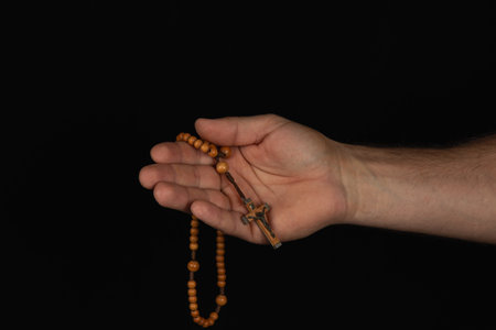 A hand is gently holding a wooden rosary with a cross, illuminated against a dark background, creating a serene and reflective atmosphere.の写真素材
