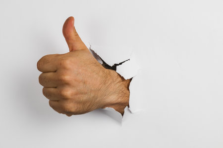 A hand emerges from a torn piece of white paper, giving a thumbs up. This image symbolizes positivity and approval in a creative way.の写真素材