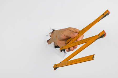 A hand emerges from a hole in the wall, gripping three yellow measuring tools. The scene showcases a...