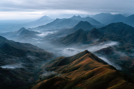 Majestic peaks stretch across the horizon, cloaked in mist and shadows. Soft clouds hover above, creating a serene atmosphere during the early morning light.の素材