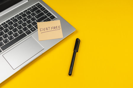 A reminder note saying debt free rests on a silver laptop beside a black pen, all set against a...