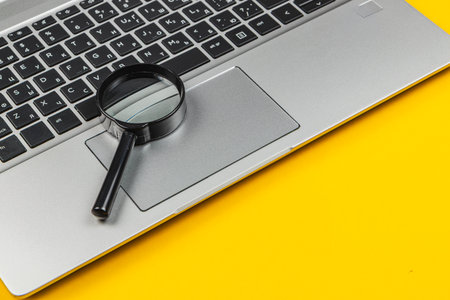 A magnifying glass rests on the touchpad of a silver laptop with a vibrant yellow background, suggesting research or detailed examination of online materials.の写真素材