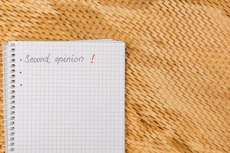 A blank checkered notebook with the phrase Second opinion! written in red ink rests on a woven mat. The setting suggests preparation for discussion or decision-making.の写真素材