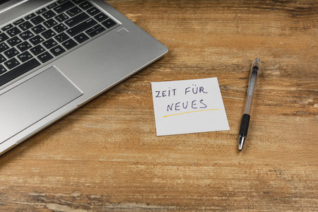 A handwritten note saying Zeit fÃ¼r Neues sits on a wooden desk beside a laptop and a pen, suggesting motivation and fresh ideas for the day.の写真素材
