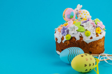 A cake sits on a blue background, decorated with flowers, candy, and small eggs. Nearby, colorful...