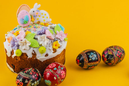 A cake topped with icing, candy, and flowers sits on a bright yellow surface. Nearby, decorated Easter eggs add to the festive scene.の写真素材