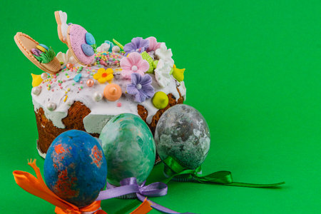 Easter cake sits next to decorated eggs. The cake has icing and colorful decorations. The eggs are...
