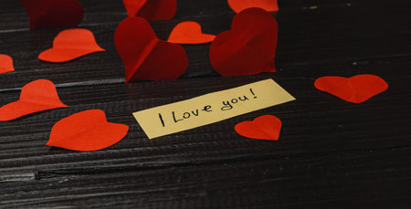 Red paper hearts scatter across a dark table, with a note saying I love you in the center. The scene...