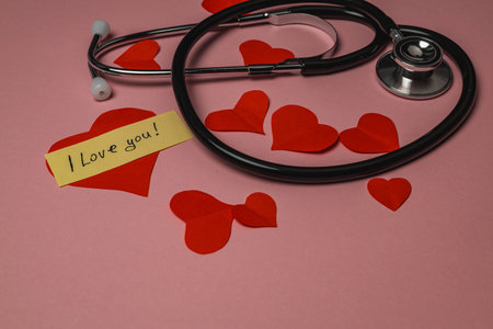 A stethoscope rests on a pink surface surrounded by red paper hearts and a note saying I love you....
