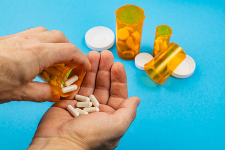A person holds a pill bottle and pours white capsules into their hand. Several other pill bottles are in the background on a blue surface.の写真素材