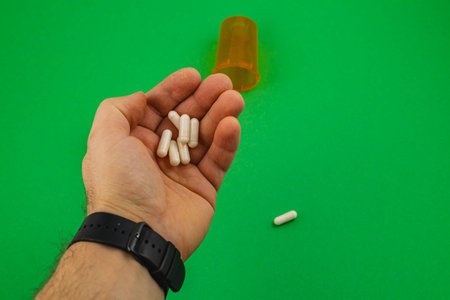 A hand holds several white capsules above a green surface. An orange pill bottle sits nearby,...