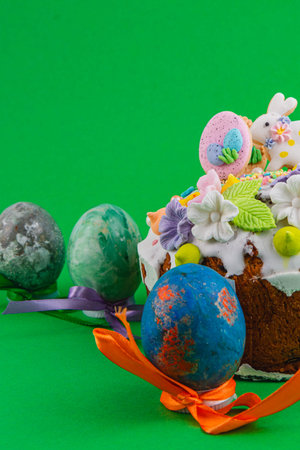A cake sits on a green background with decorations on top. Nearby are colorful eggs tied with...