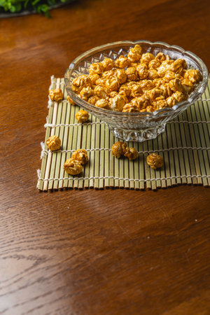 A clear bowl holds popcorn on a wooden table. Some popcorn pieces are scattered around. This scene...