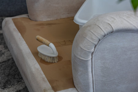 A person cleans a couch using a small brush. The couch is light in color and some dirt is visible on the surface. This cleaning happens inside a living room.の写真素材