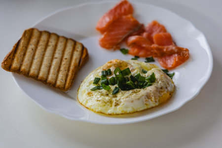 The meal shows scrambled eggs with green onions, slices of salmon, and brown toast. It is served on...