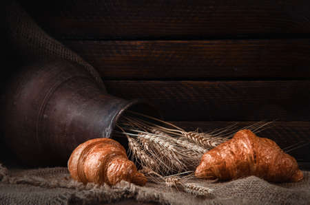 still life earthenware jug with wheat ears, bread, croissants on a drapery
