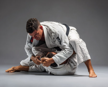 Two athletes in white gis demonstrate BJJ grappling technique in studio.の素材