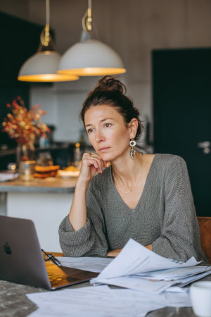 Woman deep in thought, working remotely on laptop with papers at home.の素材