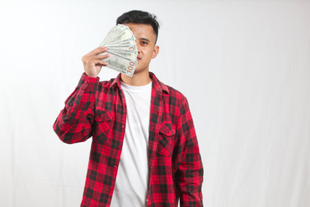 Asian man holding a hand fan with one hundred dollar bills covering half of his face isolated on white backgroundの写真素材