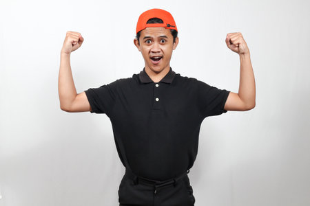 Portrait of young Asian man wearing black polo shirt and orange cap celebrating success isolated on...