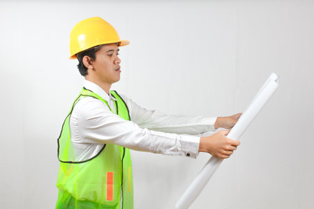 Asian worker in hard hat and safety vest with blueprint on white background