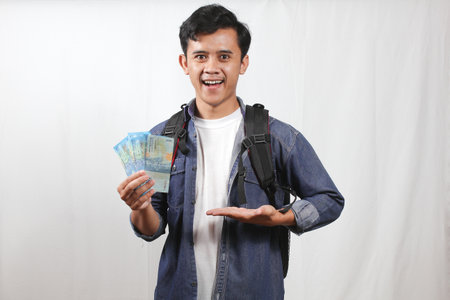 Young asian man with travel bag and holding money on white backgroundの写真素材