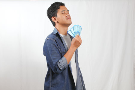 An attractive young man is enjoying the scent of a fan of money, wearing a denim jacket and white...