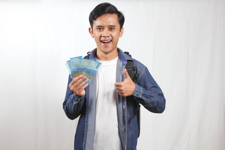 young adult man smiling and holding a fan of Indonesian Rupiah banknotes, giving a thumbs up...