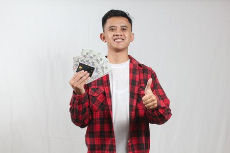 Young asian man holding money and showing thumbs up on white background