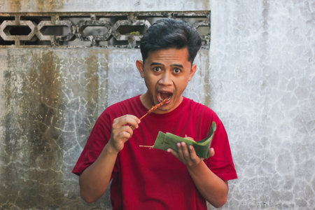 Asian man wearing a red t-shirt eating chicken satay and rice cake in the morningの写真素材