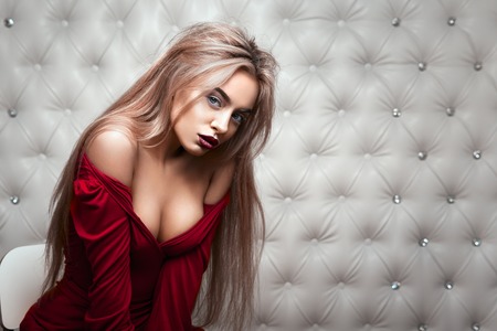 Studio portrait of a sexy blond in red dress, over leather upholstery background. Elegant luxury...