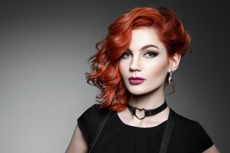 Portrait of beautiful woman with red hair and elegant hairstyle in black dress posing on gray background in studio. Fashion photo.の写真素材