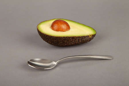 A half of an avocado with a bone and a spoon lie on a gray background.の写真素材