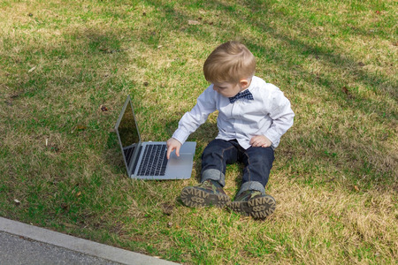 Boy is sitting on the green grass and typing on new silver laptopの写真素材