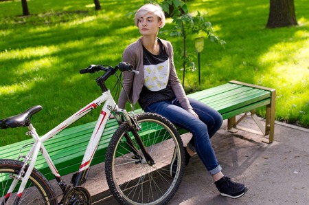 Young beautiful girl sitting on the bench near the white bicycle in the parkの写真素材