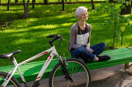 Young beautiful girl sitting on the bench near the white bicycle in the parkの写真素材