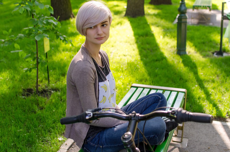 Young beautiful girl sitting on the bench near the white bicycle in the parkの写真素材