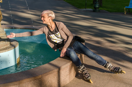 Young girl sitting near the fountain and wearing black rollers in the parkの写真素材