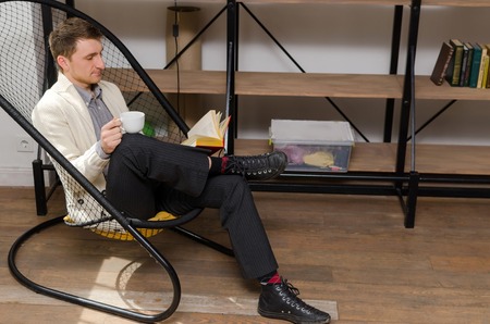 Young man sitting in the armchair with a cup of coffee is reading a bookの写真素材