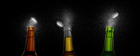 Closeup photo of an three beer bottle splashing beer drops on a black background. Beer cap flying on top of the bottleの写真素材
