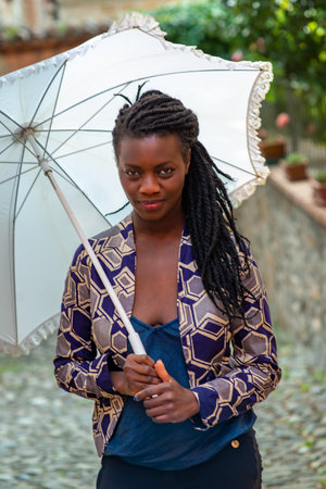 Portrait of an African woman with dreadlocks braid with umbrella parasol. Happy young woman feeling confident in her style. Fashionable woman standing in the street of old village against stone wall.の写真素材