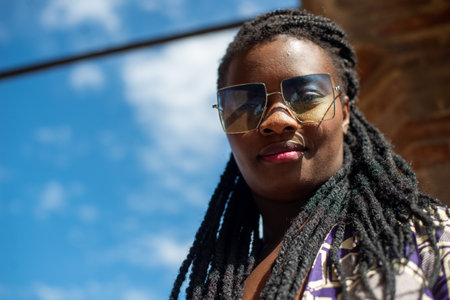 Happy young african woman feeling confident in her style. Fashionable woman wearing sunglasses and braided hairstyle outdoors. Tourist traveling italian old village.の写真素材