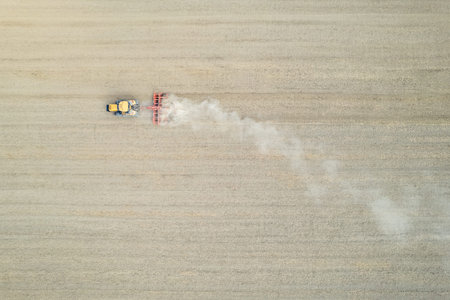 Farmer driving modern yellow crawler tractor harrowing and leveling dry soil for next seeding in...