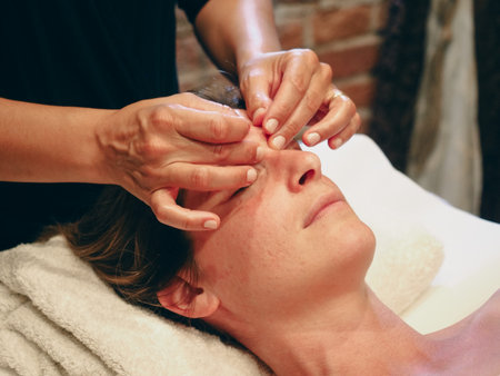 Close up hands of chiropractor or masseur making relaxing face massage to lying young fit woman at wellness spa cabin.の写真素材