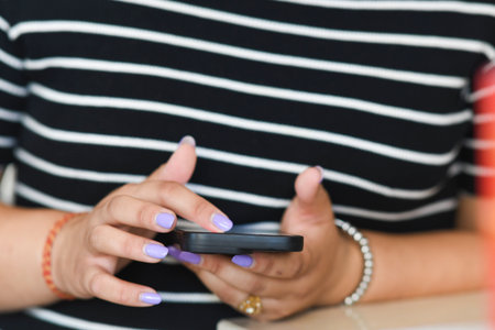 Cremona, Italy - July 21st 2025 Close-up of a woman's hands with neatly manicured nails holding and using a smartphone with a black striped shirt in the background focusing on technology andの写真素材