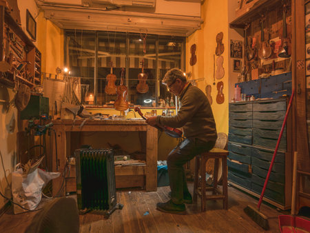 Mature adult luthier meticulously applying varnish to a violin scroll, working on handcrafted string...