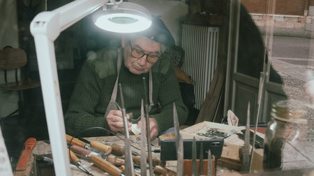 An elderly artisan with glasses focuses intently on carving a violin scroll, surrounded by his...