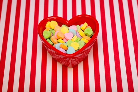 a heart shaped candy dish filled with candies is on a red and white striped background の写真素材