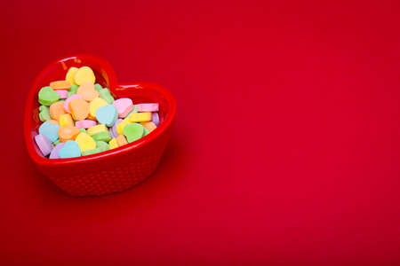 a heart shaped candy dish filled with heart candies left justified on a red textured background  copyspaceの写真素材