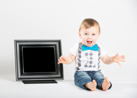 a cute 1 year old baby sits next to a blank black picture frame in a white studio setting. The boy is extremely excited and screaming. He is dressed in Tshirt, jeans, suspenders and blue bow tieの写真素材
